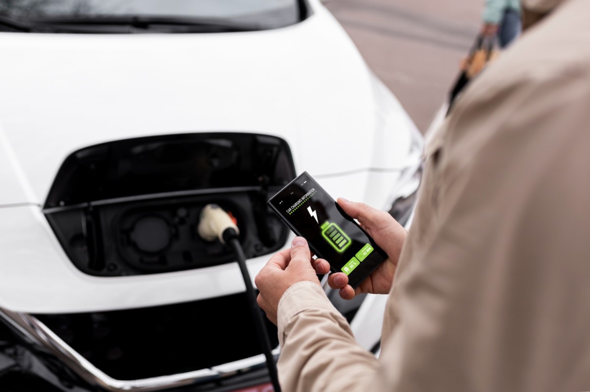 Pessoa conectando um carro elétrico branco a um carregador enquanto verifica o nível da bateria em um smartphone.