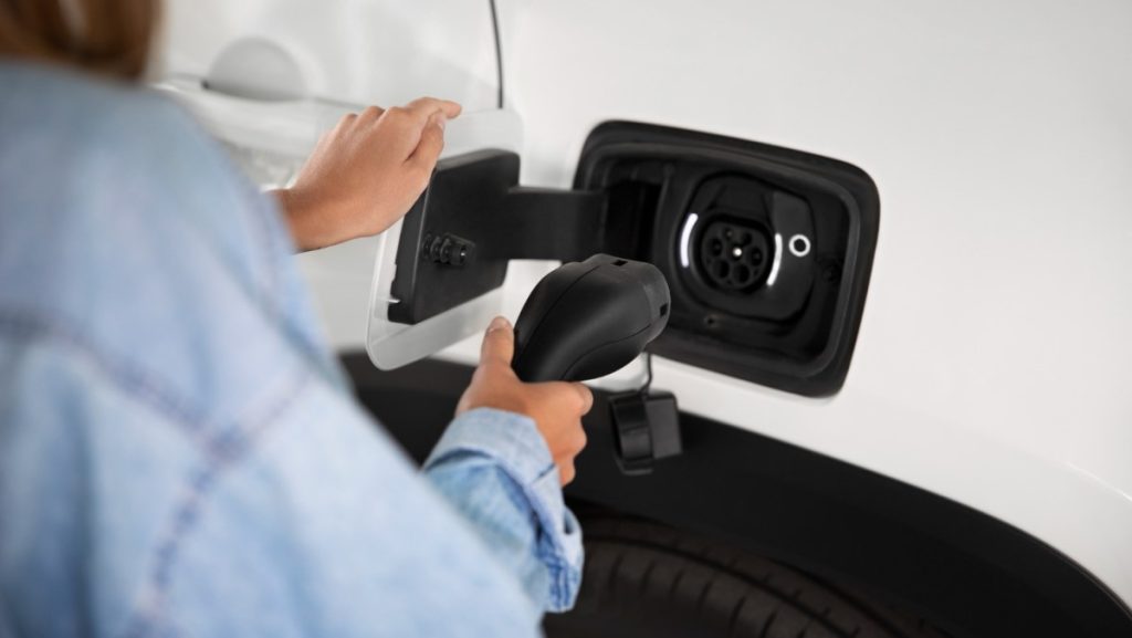 Uma pessoa conectando um carregador em um carro elétrico branco, representando a prática e a importância da mobilidade sustentável e da utilização de veículos elétricos.







