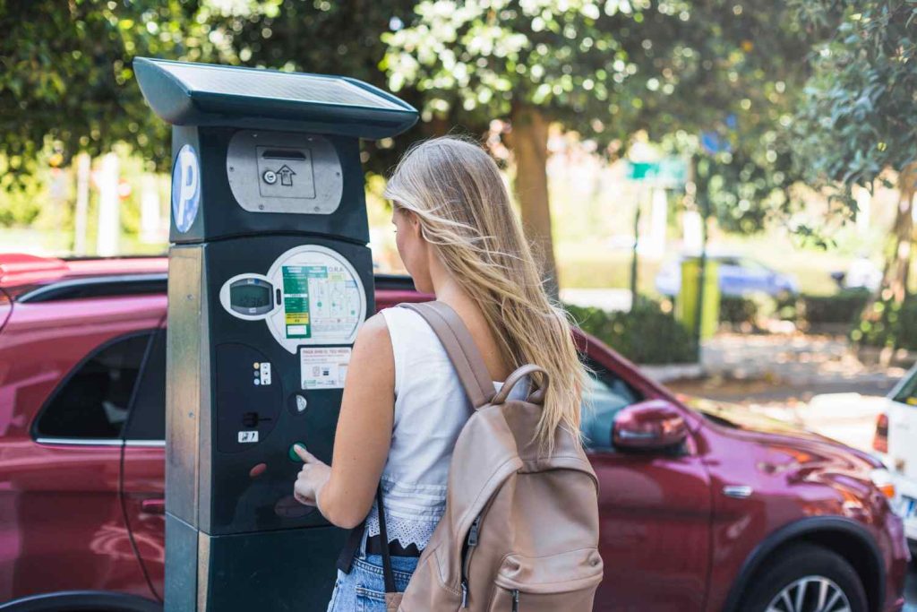 Mulher de mochila utilizando um parquímetro em uma rua com carros estacionados, simbolizando pagamento de estacionamento urbano.