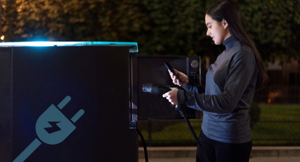 Mulher carregando um carro elétrico à noite, segurando um smartphone e conectando o carregador em uma estação de carregamento iluminada.