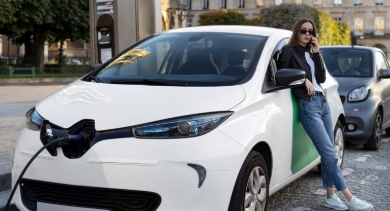 Mulher com óculos de sol apoiada em um carro elétrico branco, enquanto ele é carregado em uma estação de carregamento urbana.