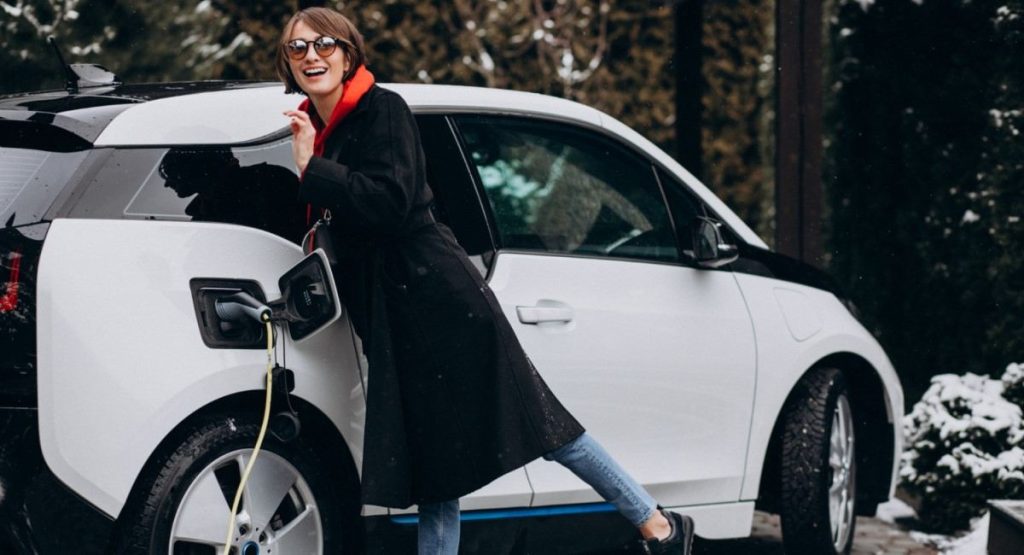 Mulher sorridente com casaco preto e cachecol laranja conectando um carro elétrico branco a um carregador em um ambiente externo.