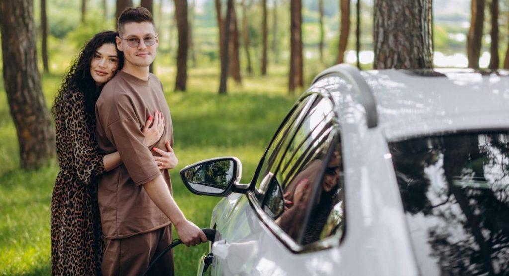 Casal jovem ao lado de um carro elétrico branco em uma área arborizada, enquanto o homem conecta o carro a um carregador.