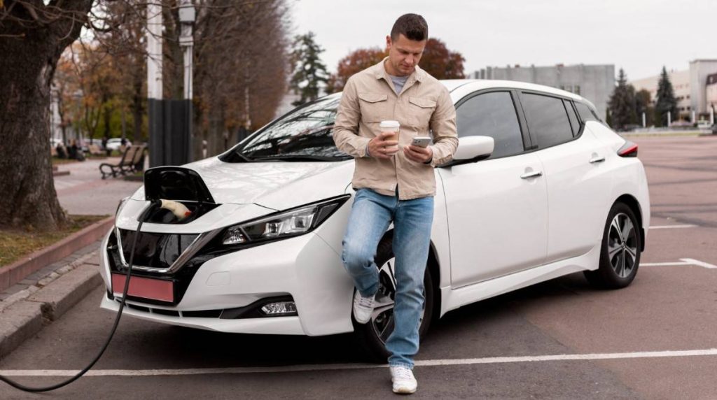 Homem encostado em um carro elétrico, segurando uma bebida e verificando seu celular enquanto o carro está carregando.