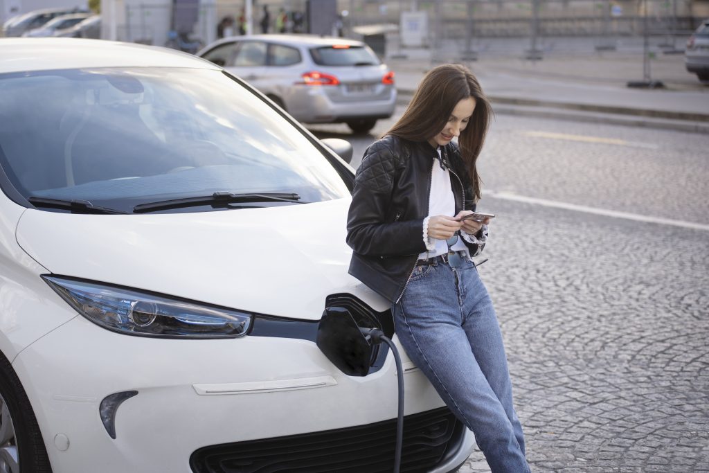 Mulher mexendo no celular apoiada no capô de um carro elétrico sendo carregado