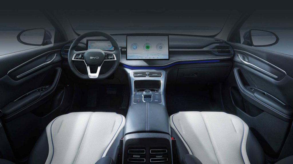 Interior do BYD King, com um painel equipado com uma tela digital que ocupa o centro, volante de formato tradicional e bancos com revestimento claro e costuras detalhadas.